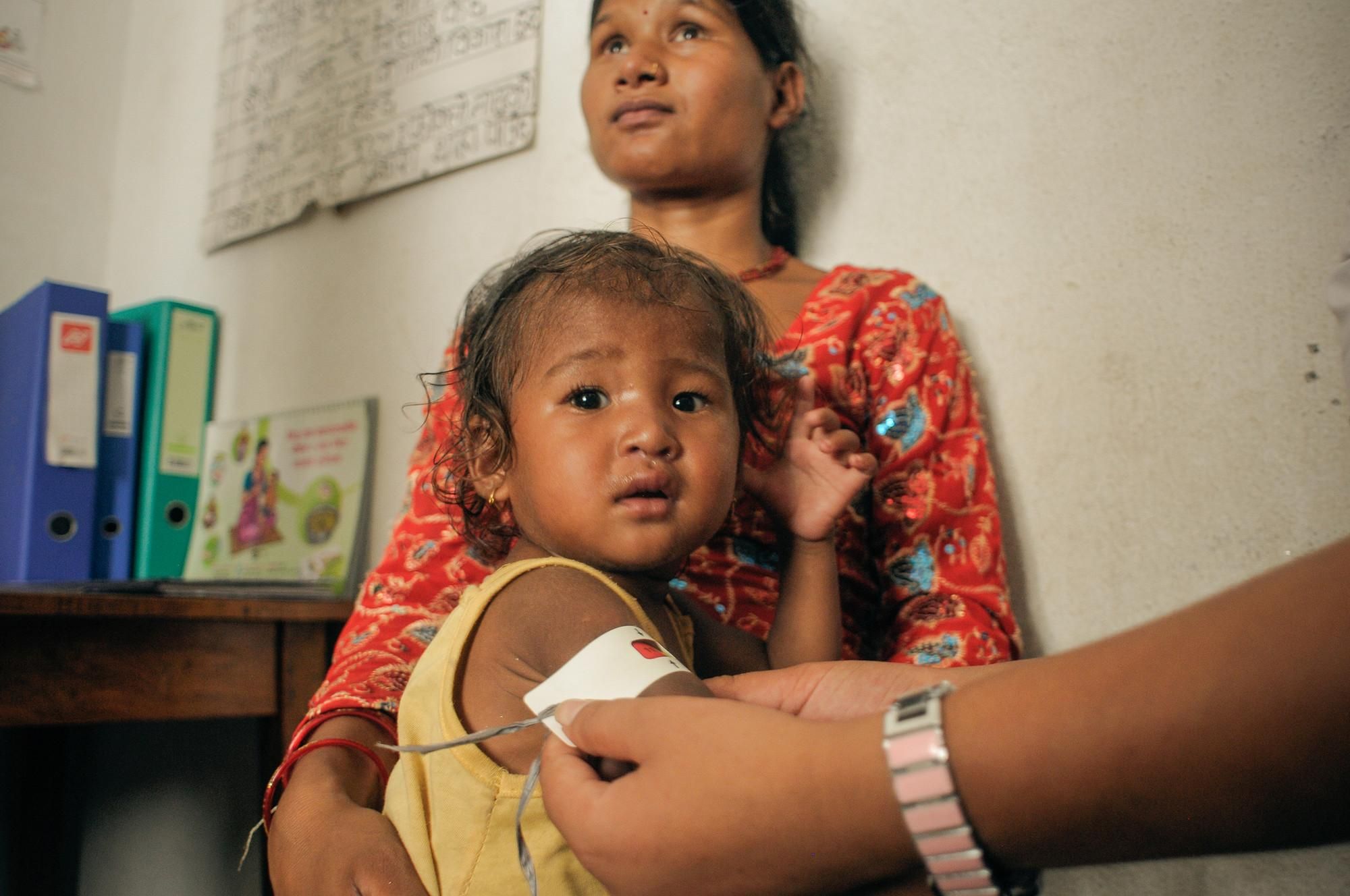 Suhila (nombre ficticio) y su madre, mientras le colocan a la niña una banda que denota que está severamente desnutrida. La pequeña fue rescatada de los escombros una hora después del terremoto. Los niños y niñas de Nepal viven en nefastas condiciones de salud e higiene. Beben agua contaminada y se ven obligados a hacer sus necesidades al aire libre.