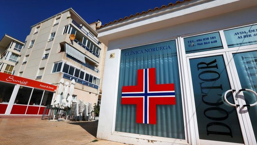 Una vecina coloca una bandera de Noruega en la ventana encima de un negocio cerrado mientras un hombre viene de comprar.