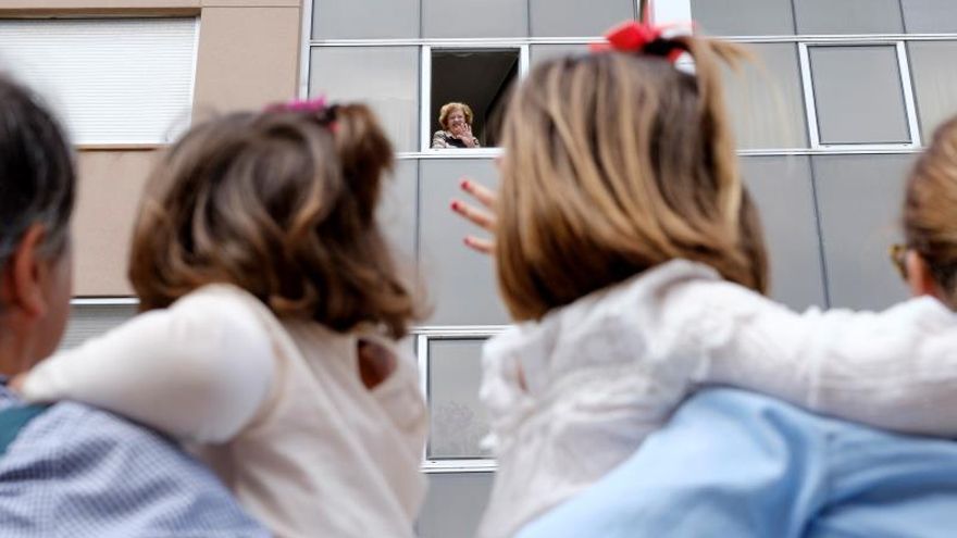 Teresa (i) y Carolina (d) aprovechan su salida a la calle para saludar a su abuela, Rosario, que les devuelve el saludo desde su ventana, esperando el día que pueda darles un abrazo.