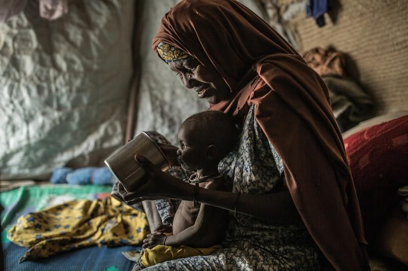 Maimuna junto a Kubi, su nieta. Tiene 50 años, huyó junto a sus hijas y nietos de la comunidad de Abuja, Nigeria, cuando Boko Haram después de la hora del rezo entró en el pueblos saqueando, matando, secuestrando y destruyendo todo. No tuvo tiempo de rescatar nada de su vivienda. Durante dos días caminó junto a su familia hasta llegar cruzar la frontera con Niger y llegar a Toumour, un pueblo que ha cuadruplicado su población los últimos años, siendo de 45.000 personas. Perdió a una de sus hijas a causa de la violencia de Boko Haram y hoy se hace cargo de sus hijos en especial de Kubi que tiene 1 año. Kubi hace dos meses que perdió a su madre y su dependencia a la leche materna la ha llevado a un deterioro crítico de su niveles de nutrición. Kubi sufre de malnutrición y su abuela le da el pecho para consolarla. El 80% de los niños y niñas de Toumour padecen de malnutrición severa. La falta de agua limpia y comida han llevado a la comunidad a una situación dramática donde muchas familias no llegan a un plato de comida diario.