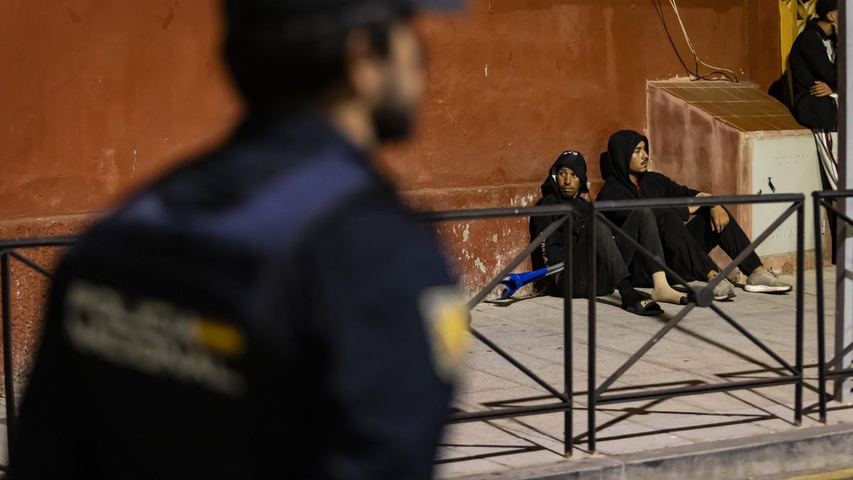 Dos personas esperan expectantes sentadas en la acera mientras los agentes van formando una línea para regular el paso hacia el puerto.