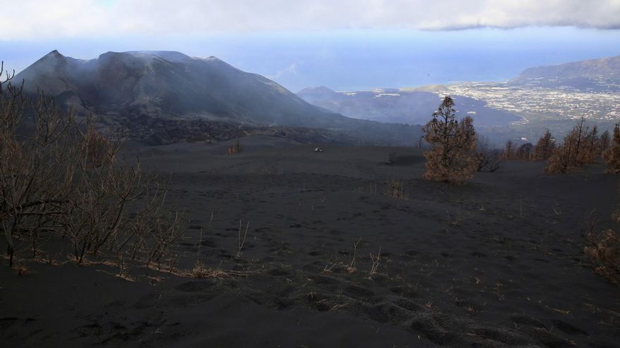 El volcán de La Palma desde Cabeza de Vaca y las coladas de lava al fondo. Cerca del edificio, se aprecia una placa solar de una estación de vigilancia volcánica y un cuarto de apero cubierto de ceniza.