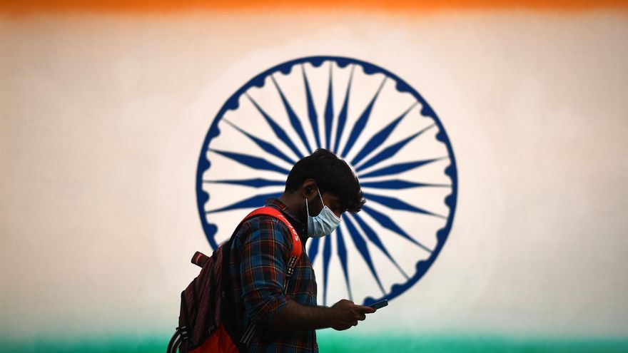 Un hombre con mascarilla revisa su teléfono móvil mientras camina frente a un mural de la bandera tricolor de la India en Chennai, India.