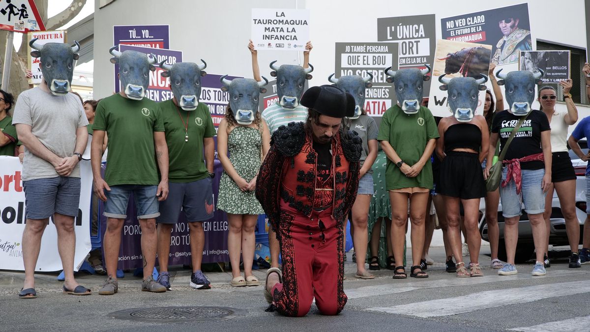 Una fila de personas con máscaras de toro mirando a una persona vestida con el traje de torero arrodillado y mirando al suelo en la manifestación antitaurina