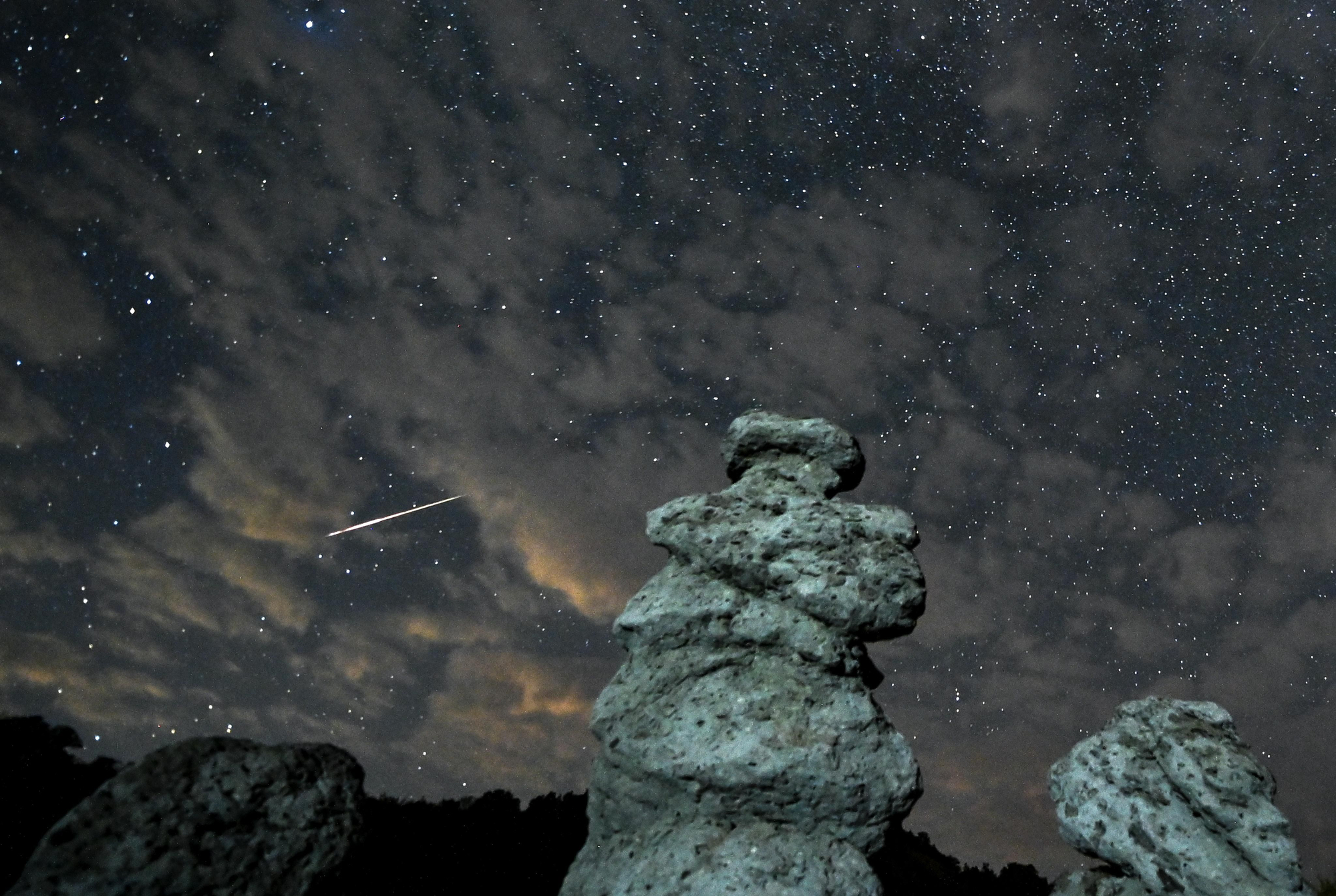 Una foto de larga exposición deja ver el rastro de una estrella fugaz que cruza el cielo nocturno entre las nubes sobre las muñecas de piedra en Kuklica, República de Macedonia del Norte, este domingo. EFE/GEORGI LICOVSKI