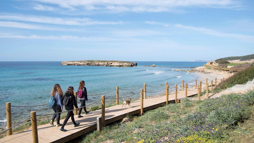 Tres jóvenes pasean por Camí de Cavalls en Menorca a su paso por la playa de Binigaus.
