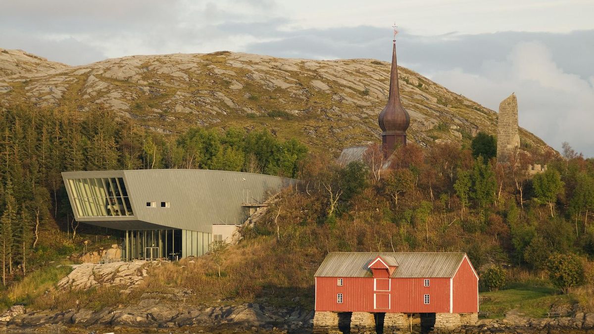 La aguja de la Iglesia de Alstahaug emerge tras los árboles. En primer término puede verse el museo dedicado a la memoria del poeta Peter Dass.