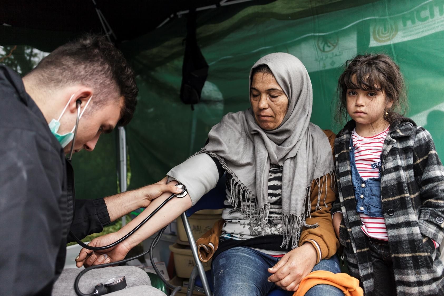 Bara, 7 años, espera mientras Momvilo Djurdjevic, doctor de MSF, toma el pulso a su tía. Proceden de Irak y tratan de llegar a Alemania, donde residen otros miembros de su familia. Muchos refugiados presentan tos,  gripe, afecciones gastrointestinales y enfermedades cutáneas, que a menudo se dan como resultado de las condiciones a las que se enfrentan durante el viaje. Los equipos también asisten a enfermos crónicos con diabetes o asma o con problemas cardíacos que no han sido atendidos de manera adecuada y que se han deteriorado en el transcurso del viaje. Fotografía: Achilleas Zavallis