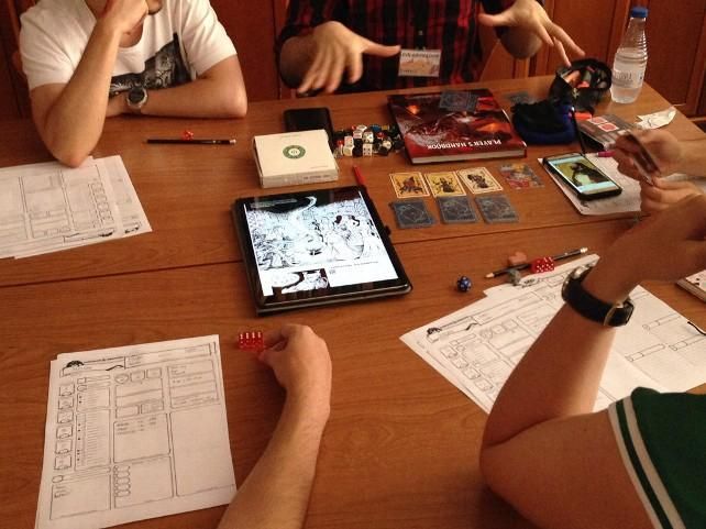 Una partida de rol de mesa, imaginando una aventura narrada con hojas de personaje en papel, cartas, dados y una tablet.