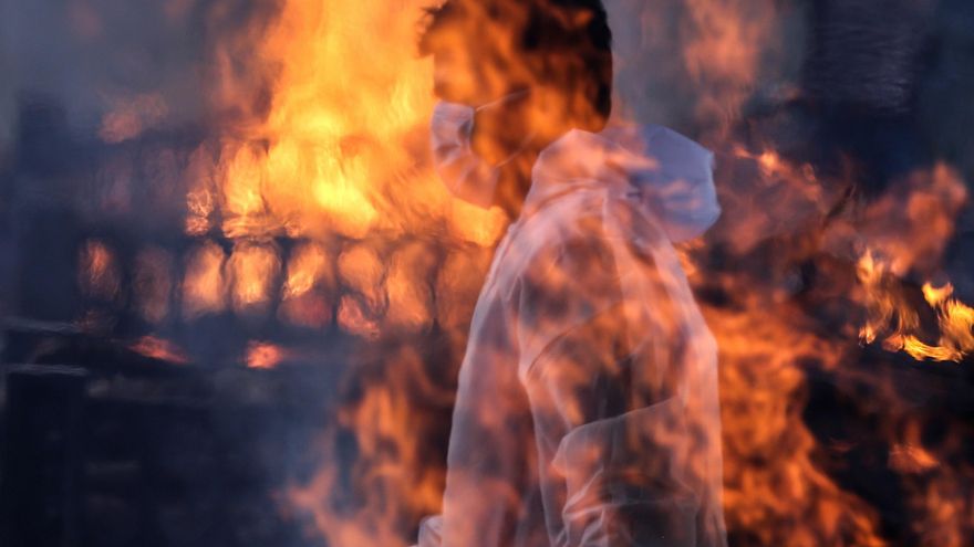 Un trabajador municipal con EPI visto tras las llamas de una de las hogueras prendidas en un crematorio para víctimas de la covid-19, este viernes en Bombay, India. EFE/ Divyakant Solanki