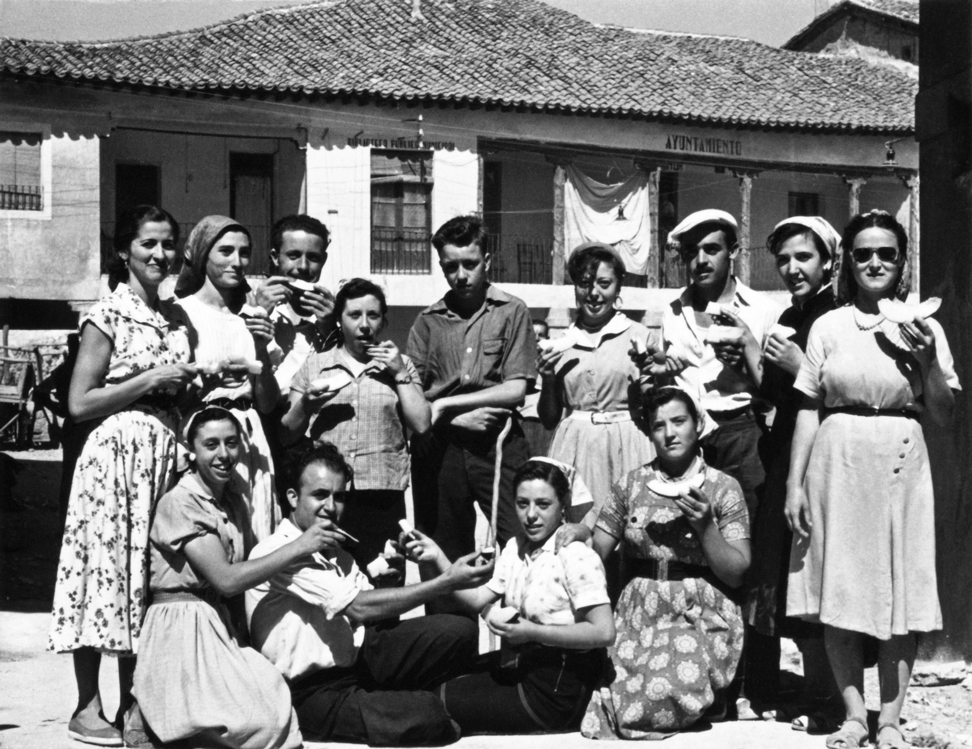Grupo de personas comiendo melón en la plaza del pueblo. Horche (Guadalajara). Años 50. Fondo Los Legados de la Tierra. Archivo de la Imagen de Castilla-La Mancha.