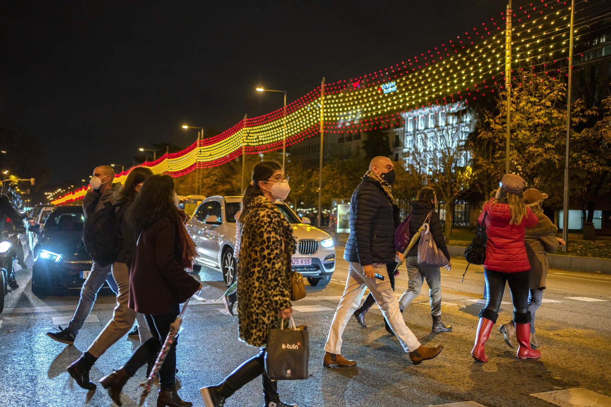 Transeúntes cruzan la calle con la bandera de luces rojas y amarillas de fondo