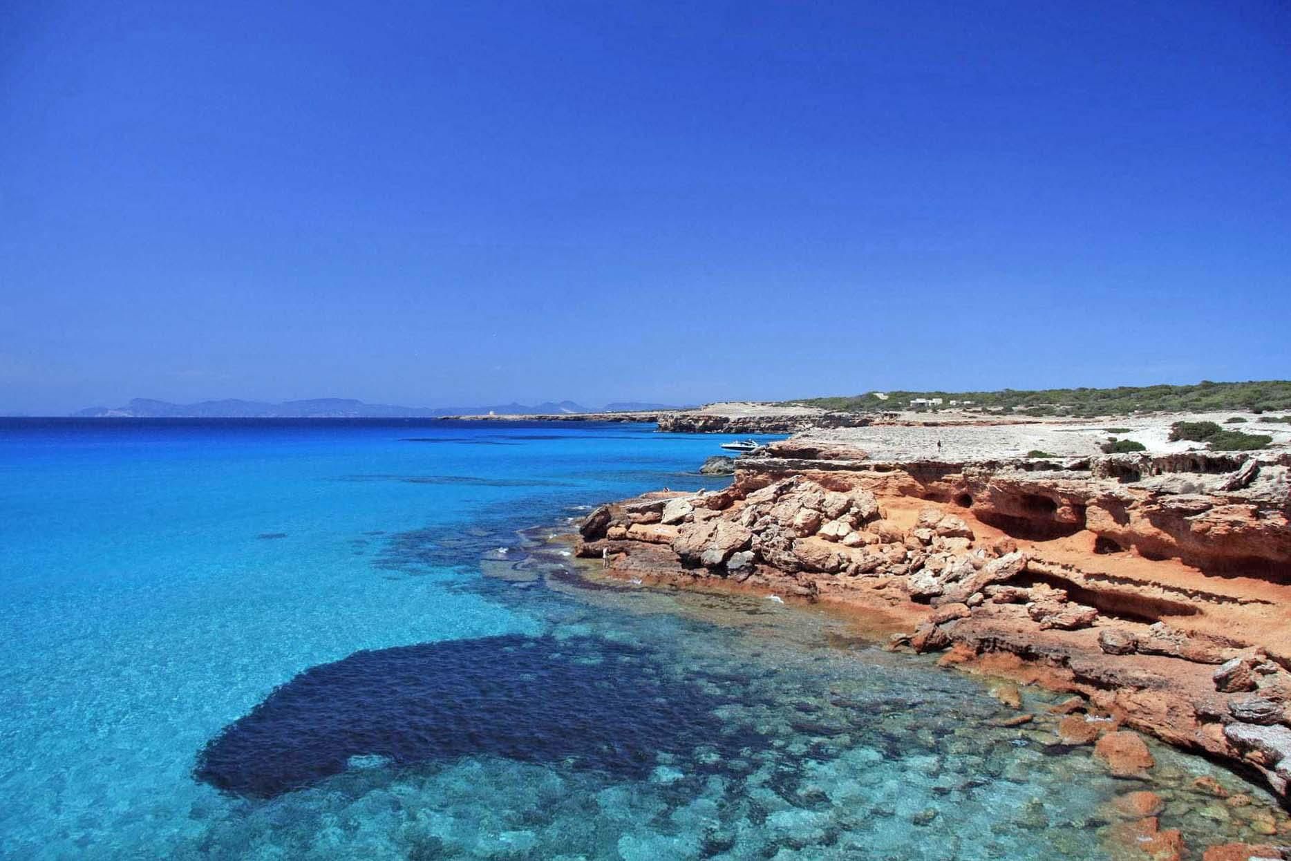 Cala Saona está rodeada por rocas de tono rojizo que bordean la playa.