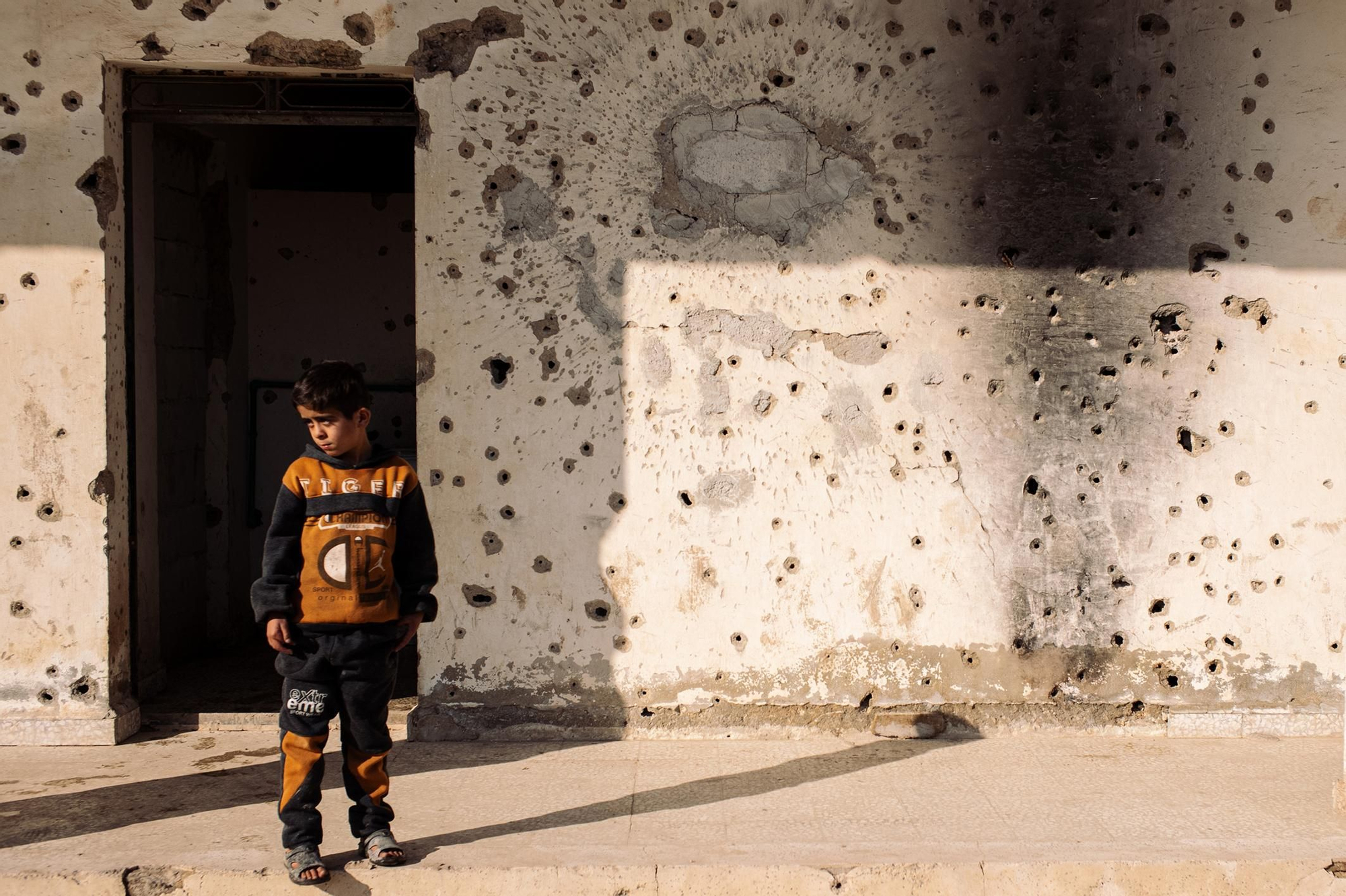 Un niño permanece frente a una pared acribillada a balazos en el campamento para personas desplazadas internas del estadio de Tabqa. Noreste de Siria, 17 de febrero de 2025. © Philémon Barbier / Hors Format. Ganador del Premio Visa d’Or Ville de Perpiñán Rémi Ochlik 2026.