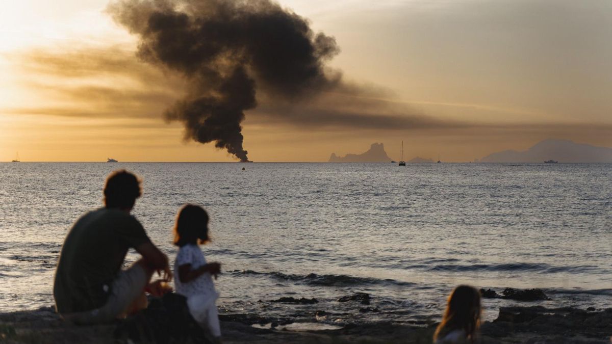 Un hombre y dos niñas observan desde la costa de Formentera el incendio en una embarcación cerca de la reserva natural de es Vedrà i es Vedranell, en Eivissa.