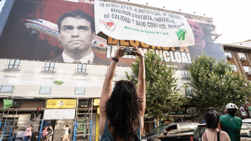 Una mujer fotografía la nueva lona desplegada por el Movimiento de Vivienda de Madrid encima del cartel de Desokupa, donde se lee ‘Ni alquileres, ni hipotecas, ni deudas. Vivienda gratuita, universal, de calidad y bajo control obrero’.