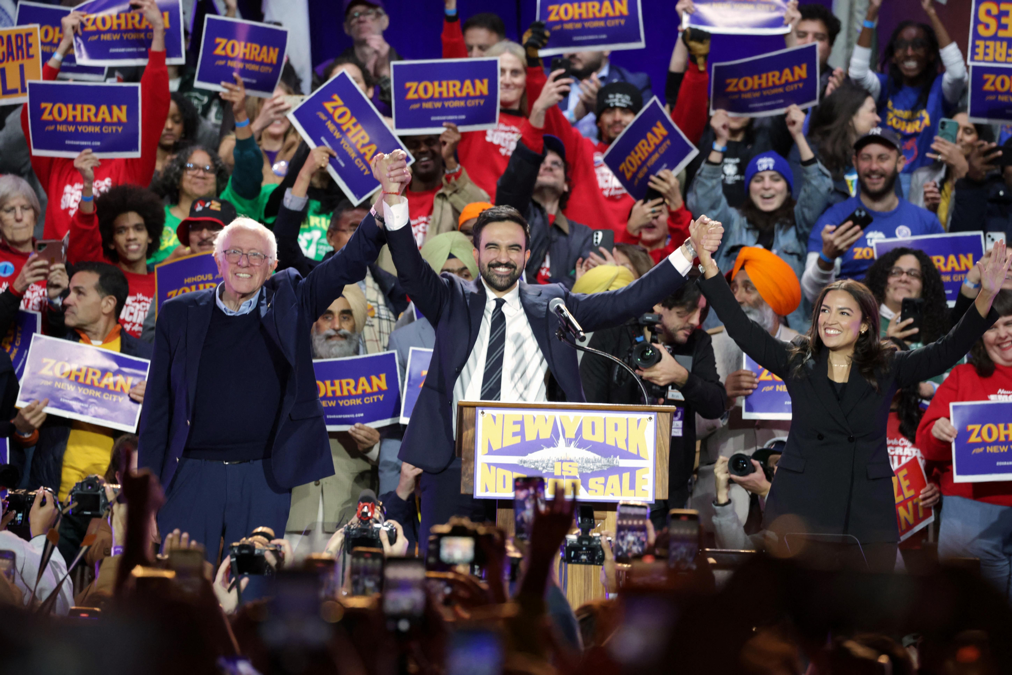 El candidato demócrata a la alcaldía de Nueva York, Zohran Mamdani, entre el senador Bernie Sanders (independiente por Vermont) y la congresista Alexandria Ocasio-Cortez (demócrata por Nueva York), al final de un acto de campaña en el estadio Forest Hills, en el distrito de Queens, Nueva York, el 26 de octubre de 2025.