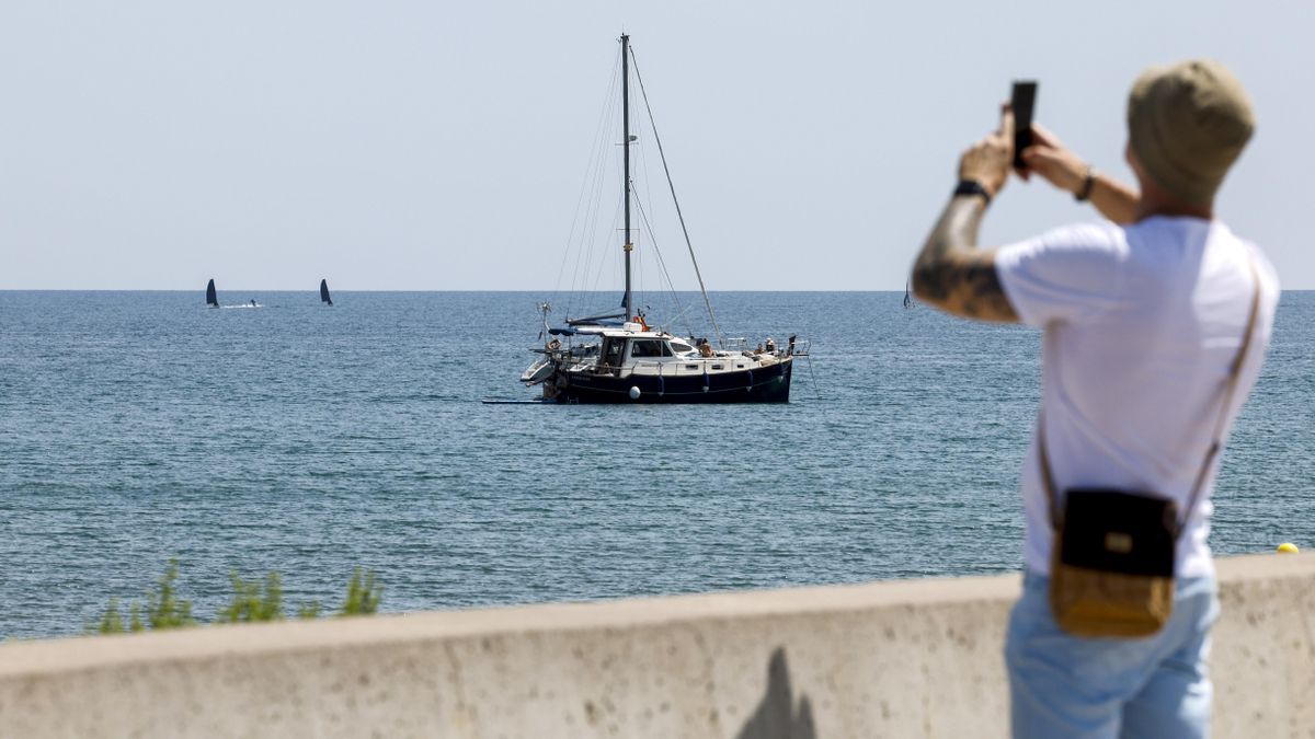 Un hombre saca una foto junto a un velero en la playa de la Malvarrosa en Valencia