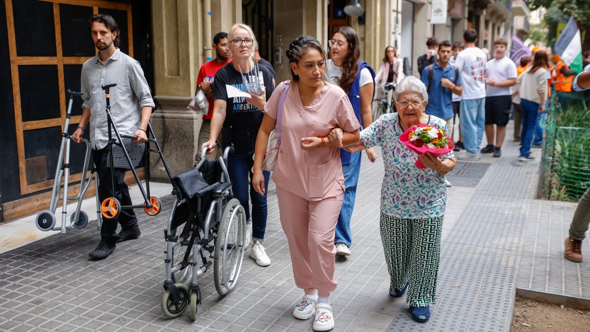 Beni, de 94 años, abandona la residencia Rosa Franch junto a una trabajadora. Al fondo, la manifestación contra la gentrificación