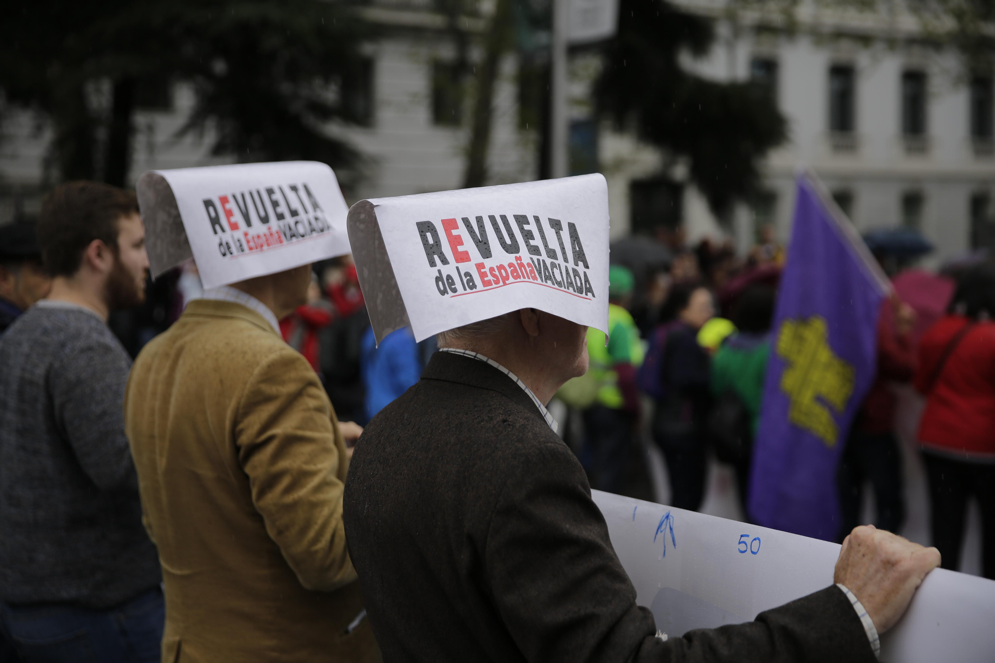 Tres asistentes a la concentración se resguardan de la lluvia con pancartas reivindicativas de la revuelta de la España vaciada