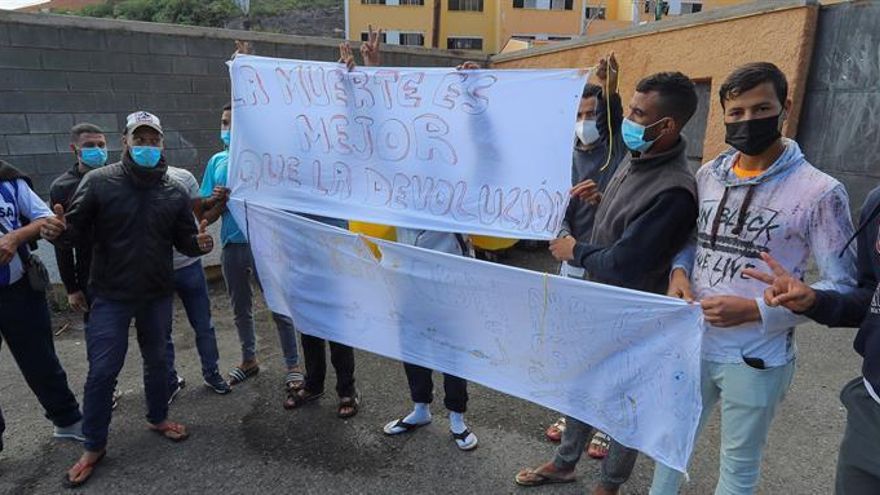varios jóvenes sostienen dos pancartas con los lemas "La muerte es mejor que la devolución" y "La migración no es un crimen" a las puertas del campamento de El Lasso