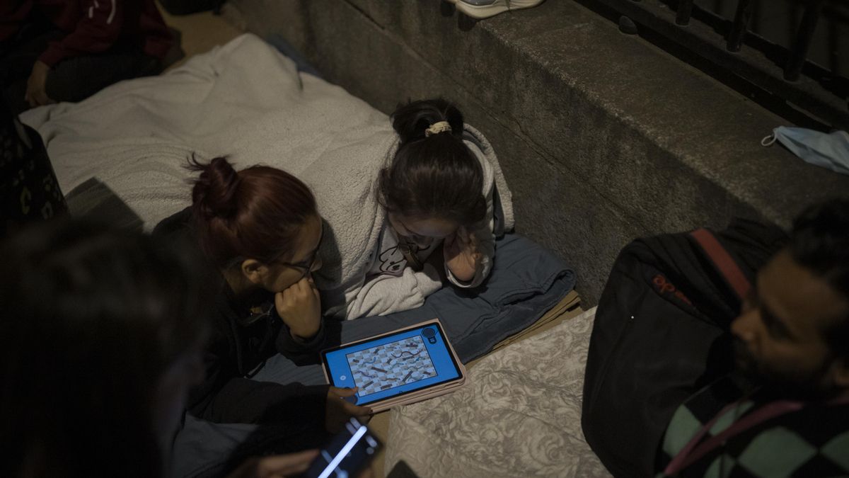 Hilary y su madre juegan con la tablet tumbadas en el suelo frente a la oficina de los servicios sociales de Madrid para pedir el informe de vulnerabilidad exigido para la regularización.