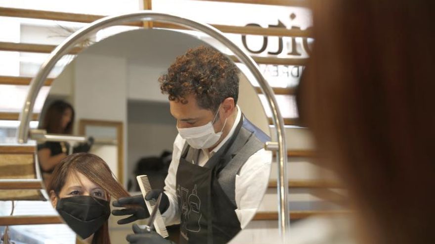 El propietario y estilista Raúl de Andreas (d) realiza un corte en su peluquería protegido con guantes y mascarilla.