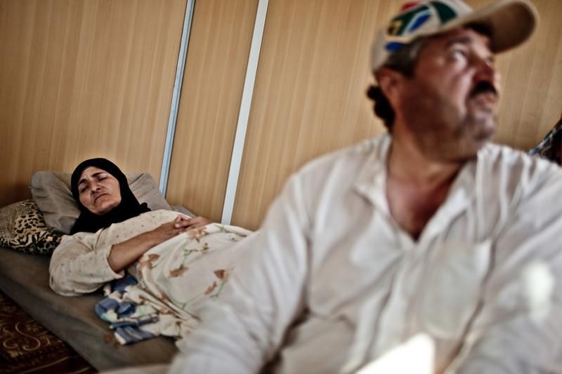 Hamda Saleem, de 49 años, y su esposo en el campo de refugiados de Za'atari, Jordania. Ella permanece tumbada en la cama. Hace meses quedó atrapada en un fuego cruzado en su ciudad, Dera’a. Recibió tres balazos, dos de ellos en el pecho –una de las balas sigue alojada ahí– y otro en la espalda. Fue tras extraerle este proyectil cuando sus piernas se paralizaron para siempre. En uno de los rincones del container su silla de ruedas acumula polvo porque es infinitamente difícil hacerla rodar sobre la tierra repleta de socavones del campo./ Pablo Tosco/Oxfam Intermón