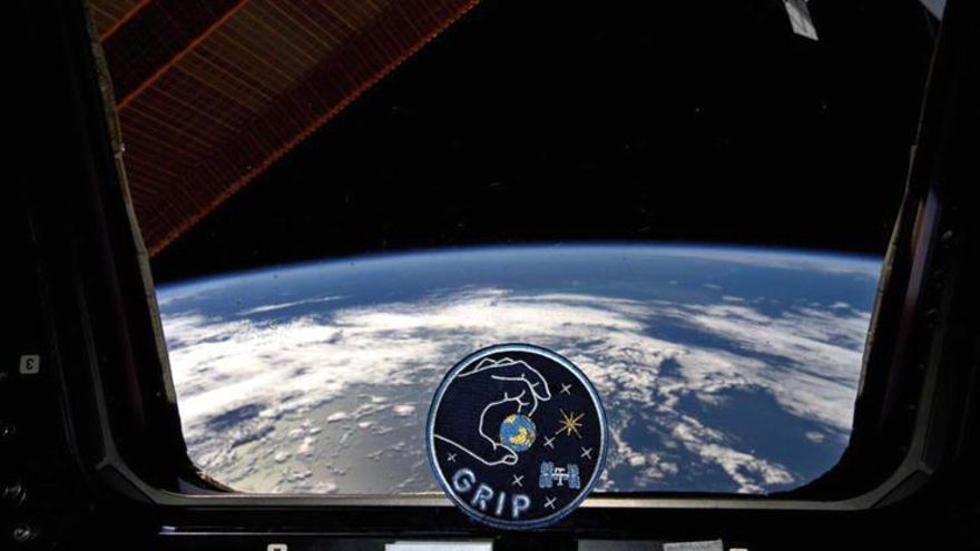 El emblema GRIP frente a la ventanilla de la ISS con la Tierra de fondo.