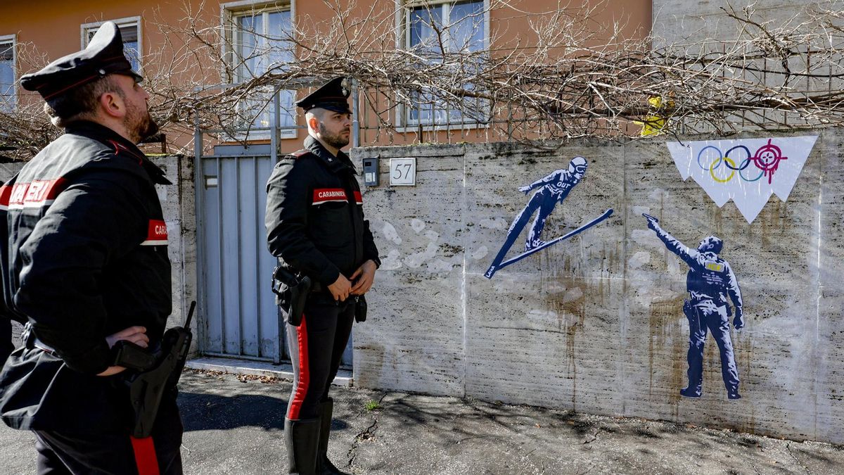Agentes de los Carabineros italianos posan frente a la nueva obra de la artista urbana Laika, titulada "¡Fuera ICE!", que representa a un agente del ICE estadounidense disparando a un saltador de esquí y una montaña volcada con el logo de los Juegos Olímpicos