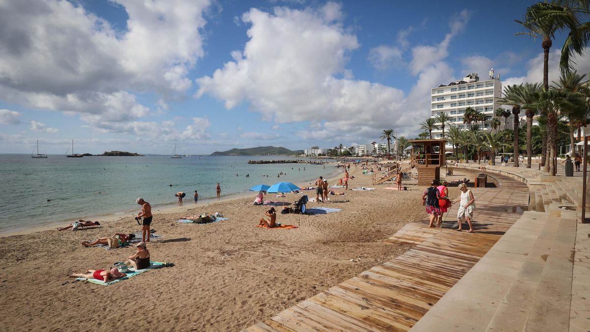 Una pasarela de madera serpentea por la arena de la playa urbana, donde destaca una torre de socorristas y varios usuarios descansan bajo un cielo soleado.