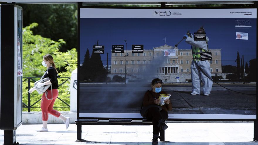 Un viajero con mascarilla se sienta en una parada de autobús frente a una imagen que forma parte de una exposición en la plaza Syntagma, en Atenas, Grecia.