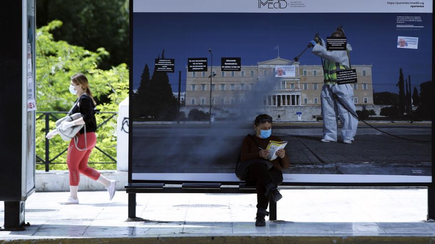 Un viajero con mascarilla se sienta en una parada de autobús frente a una imagen que forma parte de una exposición en la plaza Syntagma, en Atenas, Grecia.