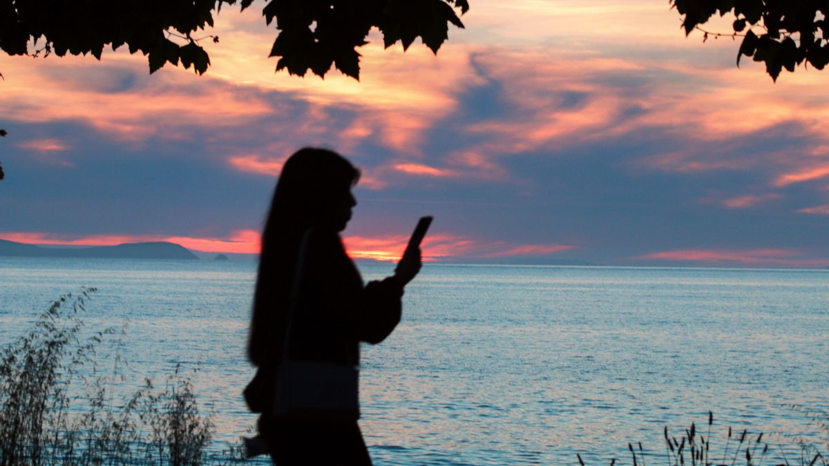 Una mujer mira la pantalla mientras cae el Sol sobre la Isla de Ons en la Ría de Pontevedra.