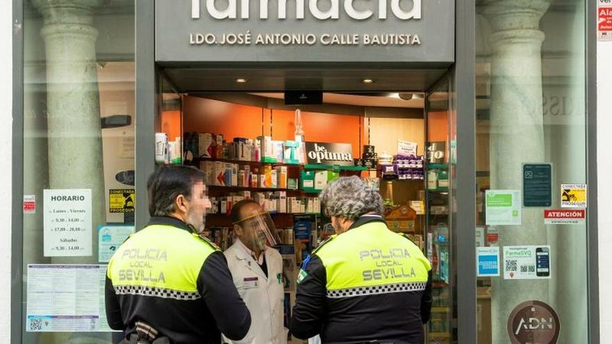 Dos agentes de la Policía Local de Sevilla hablando con un farmacéutico en la puerta de su establecimiento en la capital andaluza.