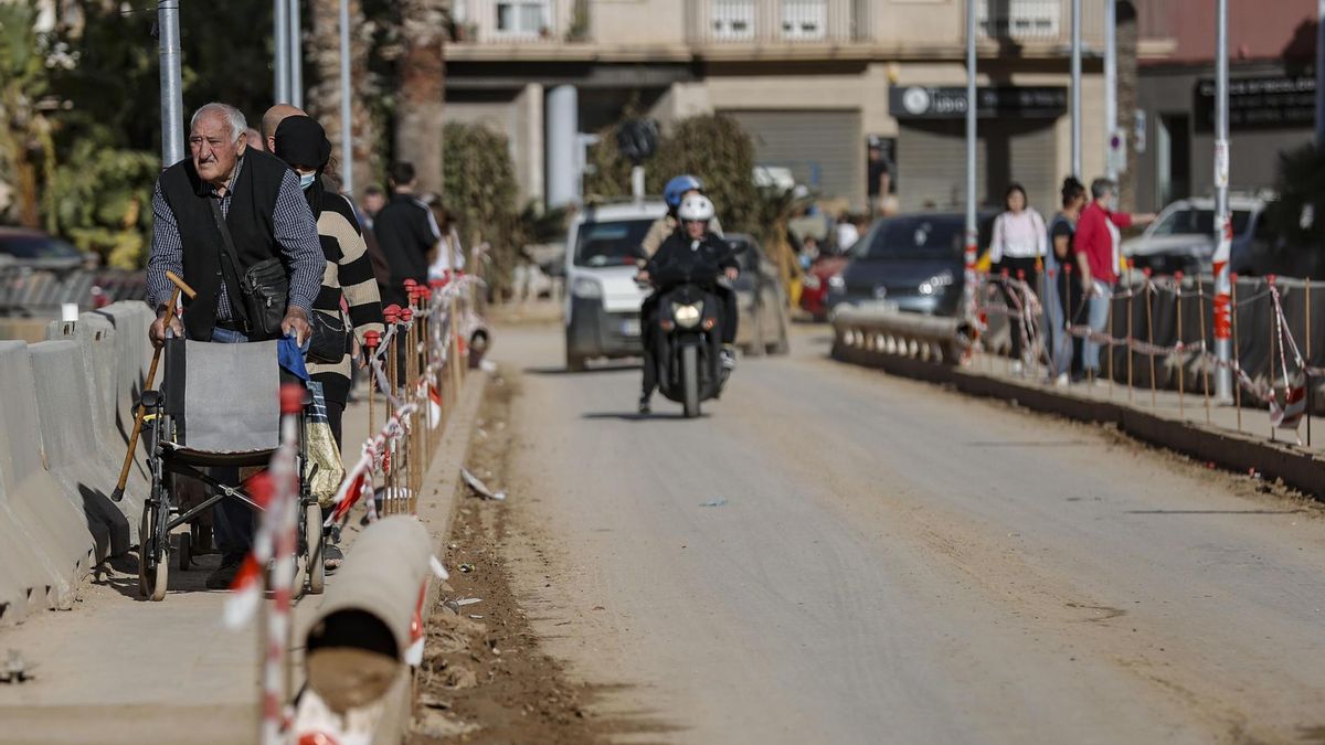 Un hombre camina apoyado en una silla de ruedas por la estrecha acera que quedó tras la dana en uno de los puentes que cruza el barranco del Poyo a su paso por Paiporta.